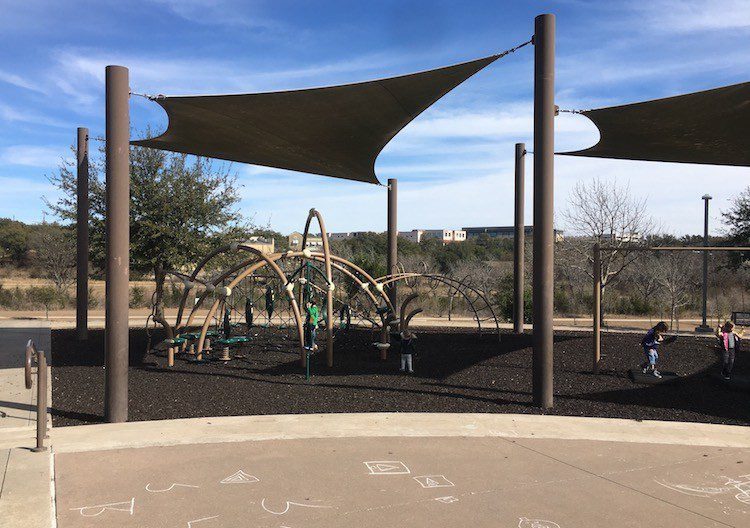 Black rubber mulch under a municipal shade-sail playground — City Playground, Bee Cave, Texas